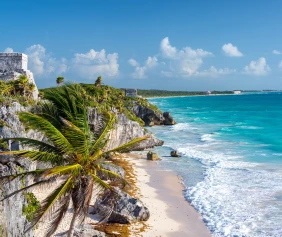 Coastal scene of Tulum, Mexico, with ancient ruins and palm trees on top of a rocky cliff, overlooking turquoise waters and a sandy beach.