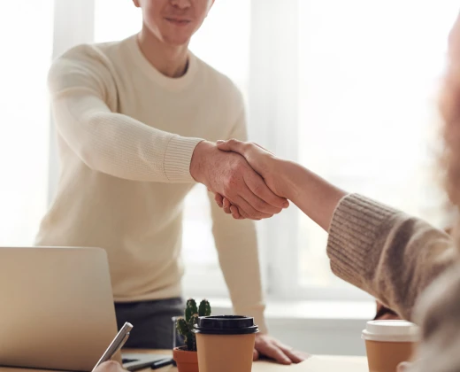 Two people shaking hands across a desk, signifying agreement or partnership. Laptop, coffee cups, and a small plant are visible, suggesting a meeting or interview setting.
