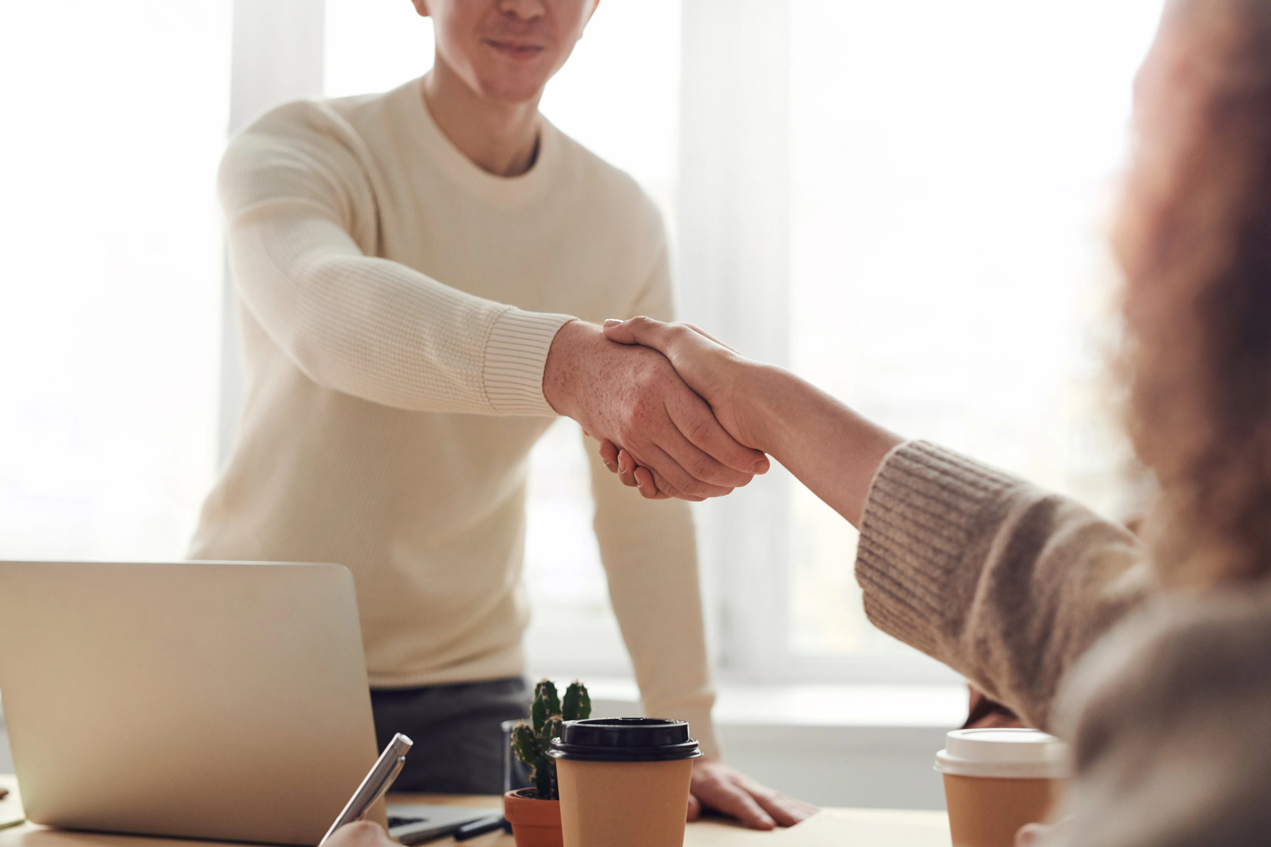 Two people shaking hands across a desk, signifying agreement or partnership. Laptop, coffee cups, and a small plant are visible, suggesting a meeting or interview setting.