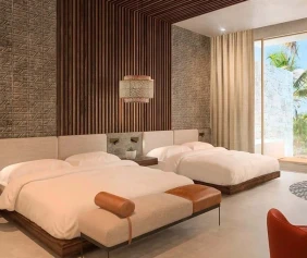 Modern hotel room with two double beds and a sleek wooden accent wall. A large window reveals a view of palm trees off the balcony.