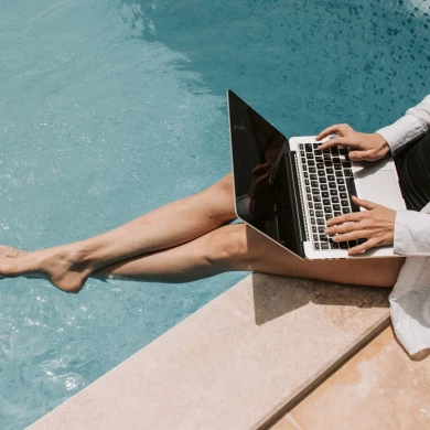 A person lounges by a poolside, working on a laptop, wearing a bathing suit with a white shirt.