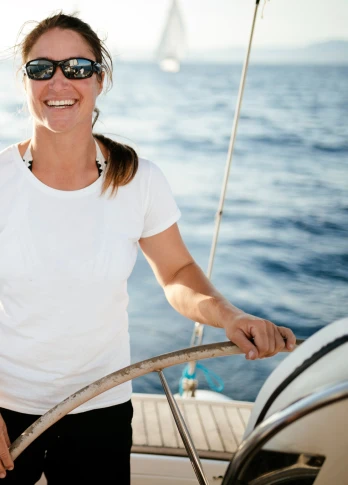 A woman in sunglasses and a white t-shirt smiles while steering a boat, surrounded by a calm sea and bright sunlight.