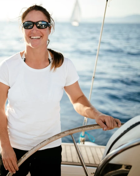 A woman in sunglasses and a white t-shirt smiles while steering a boat, surrounded by a calm sea and bright sunlight.
