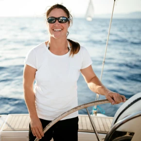 A woman in sunglasses and a white t-shirt smiles while steering a boat, surrounded by a calm sea and bright sunlight.