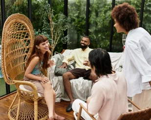 A group of four friends in casual clothing enjoy a lively conversation on a sunlit patio with plants.