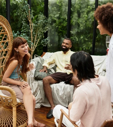 A group of four friends in casual clothing enjoy a lively conversation on a sunlit patio with plants.