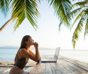 A woman seated at a wooden table outdoors, using a laptop under palm trees by the ocean. She appears thoughtful, gazing into the distance.