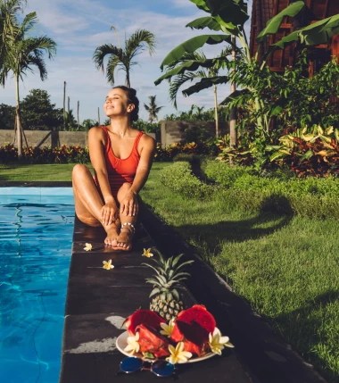 Woman in a red swimsuit sits by a pool, surrounded by greenery and palm trees, with a plate of fresh fruit in front of her.