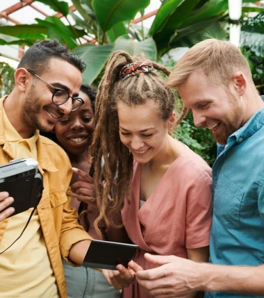 Four friends stand in a lush greenhouse, smiling and gathered around a camera and phone. They appear joyful, sharing a moment of laughter.