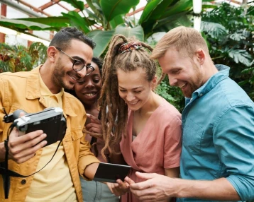 Four friends stand in a lush greenhouse, smiling and gathered around a camera and phone. They appear joyful, sharing a moment of laughter.