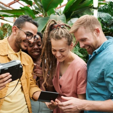 Four friends stand in a lush greenhouse, smiling and gathered around a camera and phone. They appear joyful, sharing a moment of laughter.