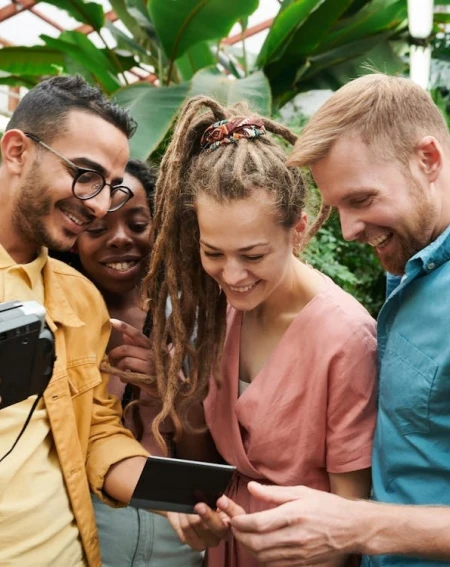 Four friends stand in a lush greenhouse, smiling and gathered around a camera and phone. They appear joyful, sharing a moment of laughter.