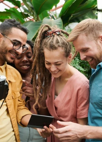 Four friends stand in a lush greenhouse, smiling and gathered around a camera and phone. They appear joyful, sharing a moment of laughter.