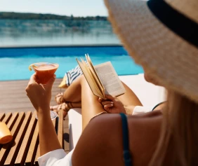 A person relaxes by a pool, lounging on a chair, holding a cocktail in one hand and a book in the other.