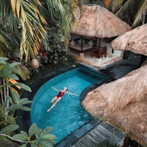 A person in a red swimsuit floats on their back in a turquoise pool surrounded by lush palm trees and tropical thatched-roof huts.