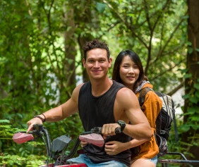 A smiling couple rides a motorized bike through a lush forest.