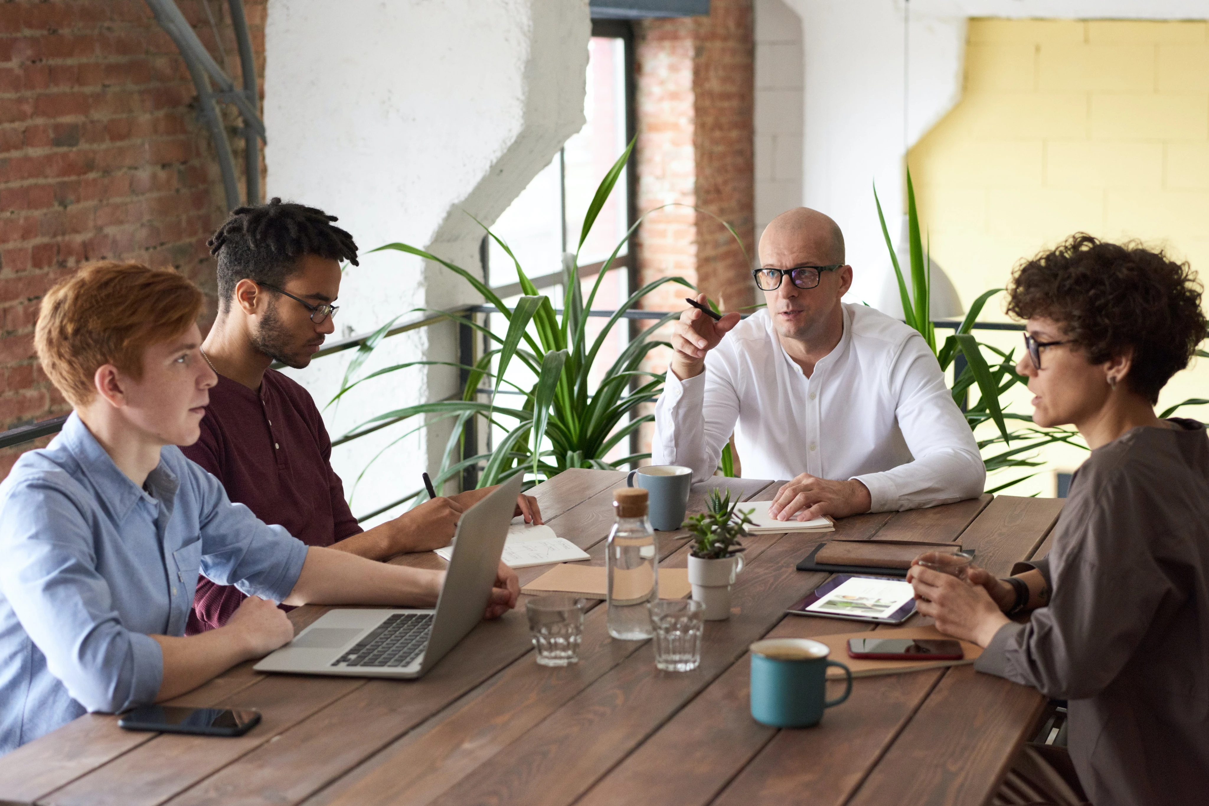 Four people in a modern office having a discussion around a wooden table. Laptops and mugs are on the table; green plants are in the background.