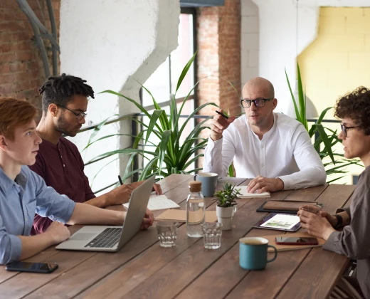 Four people in a modern office having a discussion around a wooden table. Laptops and mugs are on the table; green plants are in the background.