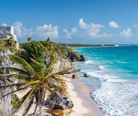 Coastal scene of Tulum, Mexico, with ancient ruins and palm trees on top of a rocky cliff, overlooking turquoise waters and a sandy beach.