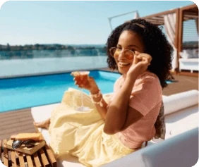 Smiling woman on a sunny poolside deck, wearing sunglasses and holding a drink. She's seated on a lounge chair, exuding relaxation and joy.