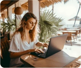 A woman with wavy hair works on a laptop at a wooden table in a tropical cafe. Sunlit palm trees and outdoor seating create a relaxed atmosphere around her.