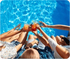 A group of people sitting by a pool raises colourful drinks together, surrounded by clear blue water.