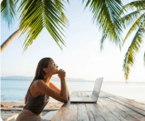 A woman seated at a wooden table outdoors, using a laptop under palm trees by the ocean. She appears thoughtful, gazing into the distance.