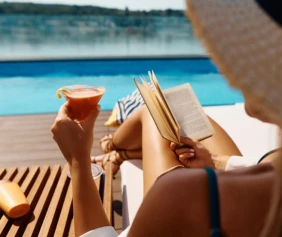 A person relaxes by a pool, lounging on a chair, holding a cocktail in one hand and a book in the other.