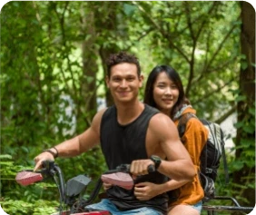 A smiling couple rides a motorized bike through a lush forest.
