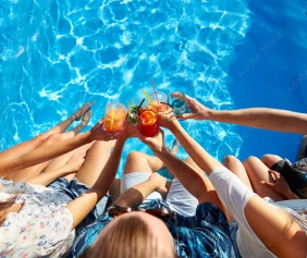 A group of people sitting by a pool raises colourful drinks together, surrounded by clear blue water.