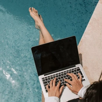 A person sits poolside, legs in water, typing on a laptop