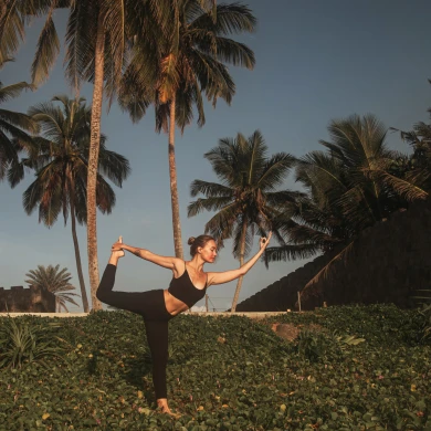 A woman practices yoga outdoors in a graceful pose, balancing on a hill. Palm trees and a clear blue sky create a serene backdrop.