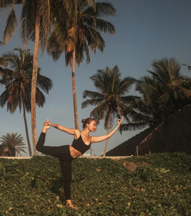 A woman practices yoga outdoors in a graceful pose, balancing on a hill. Palm trees and a clear blue sky create a serene backdrop.