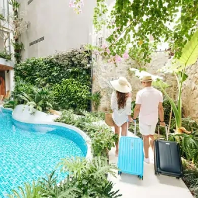A couple in summer attire and hats, each with luggage, walks beside a lush pool surrounded by greenery and stone walls, creating a serene vacation vibe.
