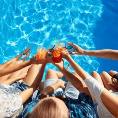 A group of people sitting by a pool raises colourful drinks together, surrounded by clear blue water.