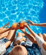 A group of people sitting by a pool raises colourful drinks together, surrounded by clear blue water.