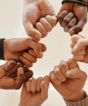 Eight diverse hands forming a circle with pinky fingers interlocked. The image conveys unity and teamwork against a neutral background.