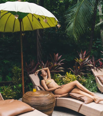 A woman relaxes on a sun lounger under a green umbrella in a tropical garden, surrounded by lush plants.