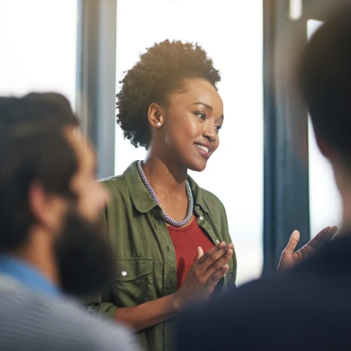 A woman in a green jacket speaks confidently to a group, smiling and gesturing with her hands. Blurred audience members listen attentively.