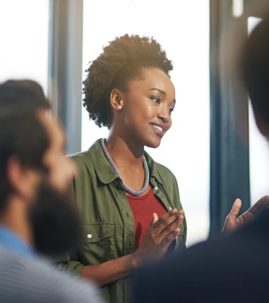A woman in a green jacket speaks confidently to a group, smiling and gesturing with her hands. Blurred audience members listen attentively.