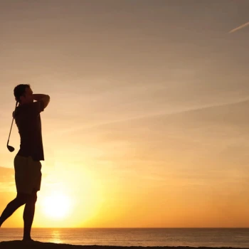Silhouette of a man golfing at sunset over the ocean, with the sky a bright orange and yellow.