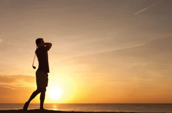 Silhouette of a man golfing at sunset over the ocean, with the sky a bright orange and yellow.