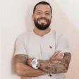 A man with tattoos and a beard smiles warmly with arms crossed. He's wearing a light shirt and a white watch, standing against a soft pink background.