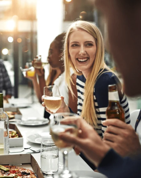 A group of friends enjoys an outdoor gathering. A woman in a striped shirt smiles, holding a wine glass. Drinks and pizza on the table create a lively atmosphere.