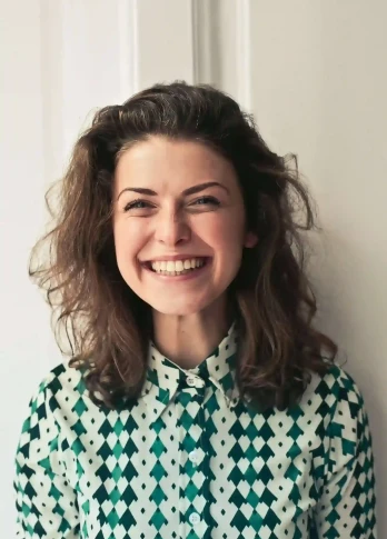 Smiling woman with wavy hair in a green and white patterned shirt stands against a white background.