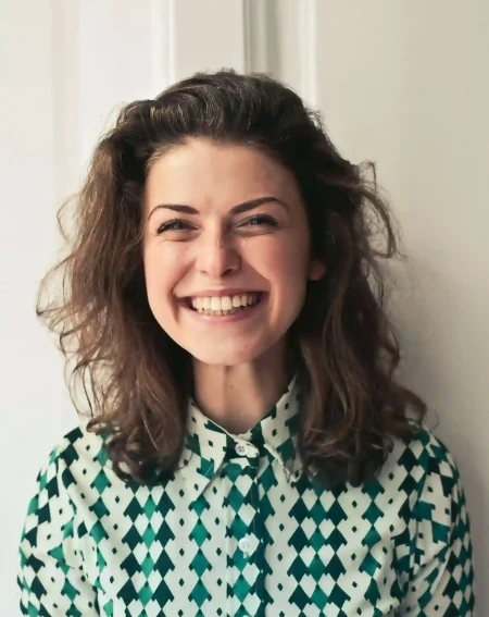 Smiling woman with wavy hair in a green and white patterned shirt stands against a white background.