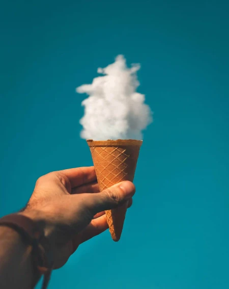 Hand holding an ice cream cone filled with fluffy white clouds against a clear blue sky, creating a surreal effect.