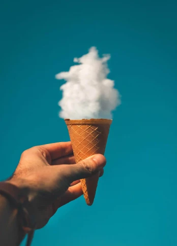 Hand holding an ice cream cone filled with fluffy white clouds against a clear blue sky, creating a surreal effect.