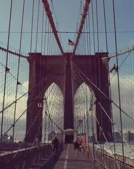 View of the Brooklyn Bridge with its iconic cables framing the path. A few people walk along the pedestrian lane. The sky is partly cloudy.