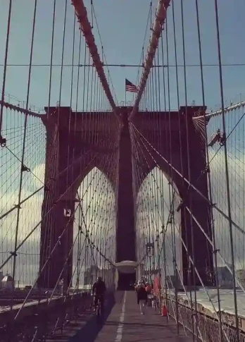 View of the Brooklyn Bridge with its iconic cables framing the path. A few people walk along the pedestrian lane. The sky is partly cloudy.